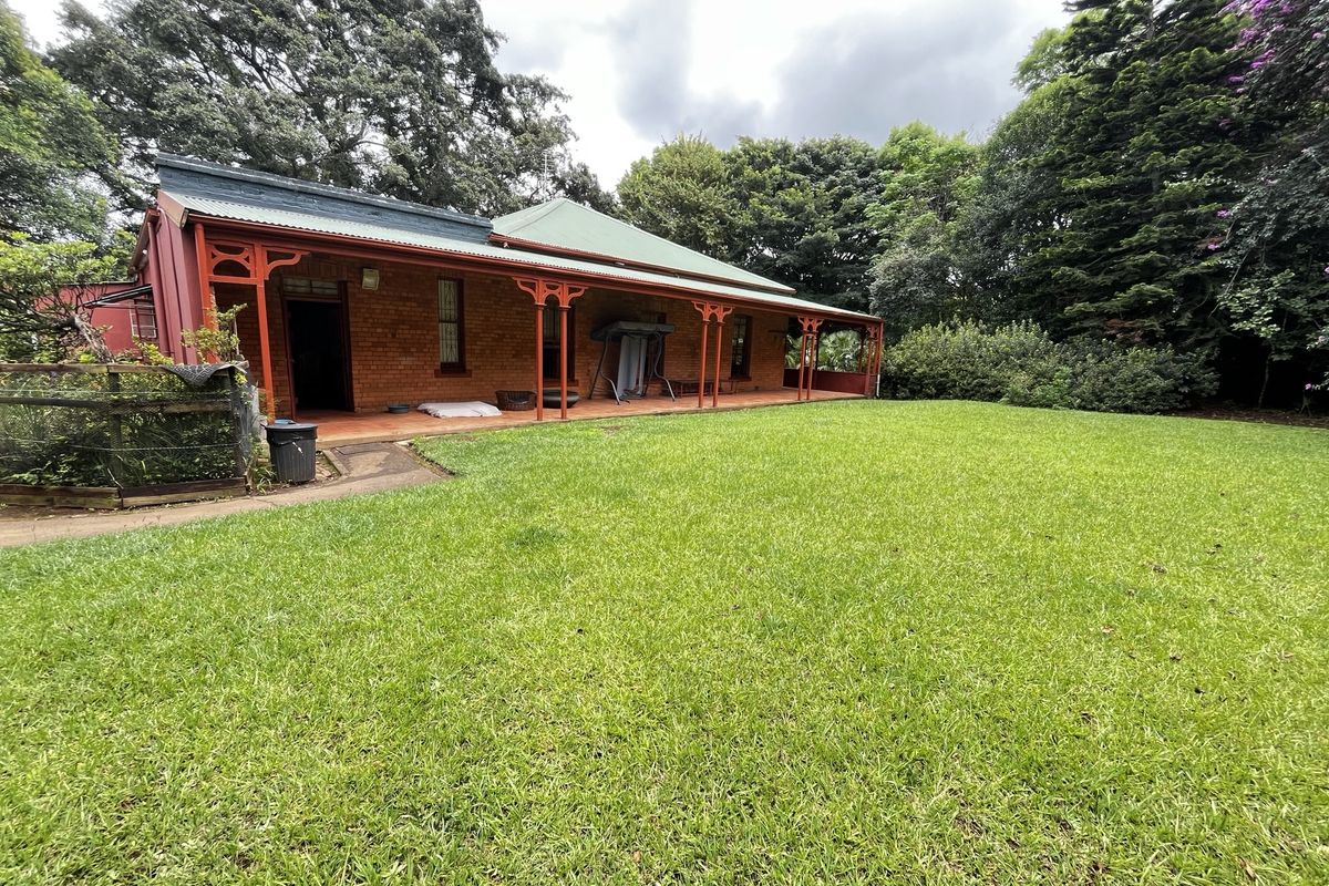 A rear view of a traditional brick house featuring a covered veranda, expansive lawn, and mature garden surroundings.