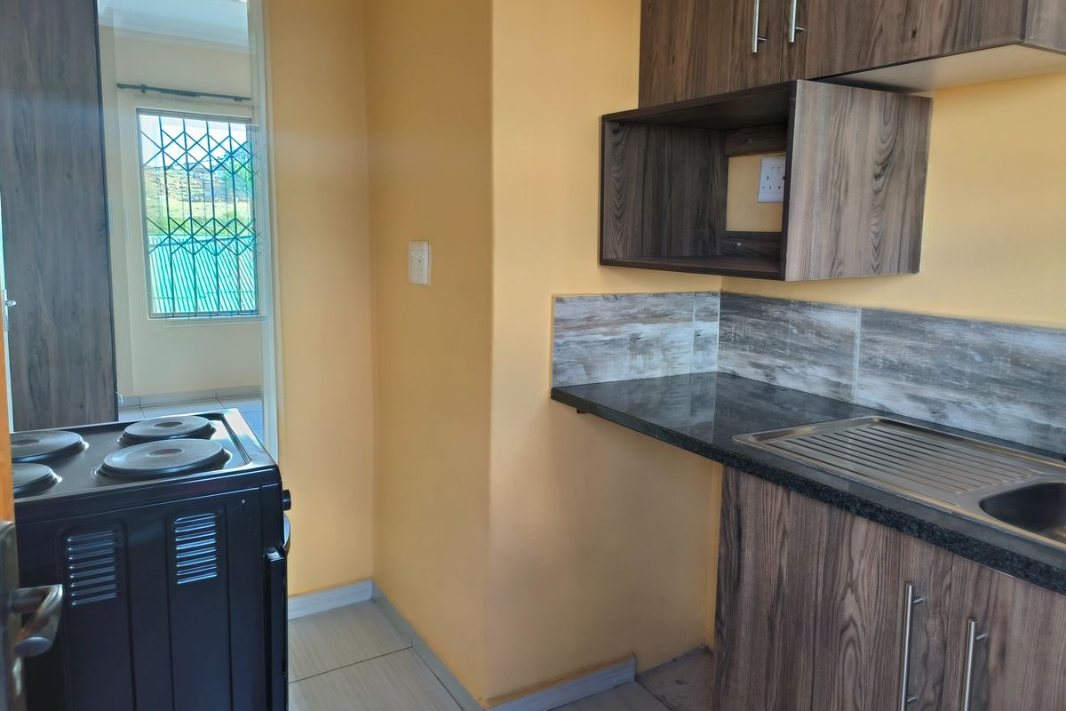 A compact kitchen featuring wood-look cabinetry, dark countertops, a stainless steel sink, and a freestanding stove, with tiled flooring throughout.
