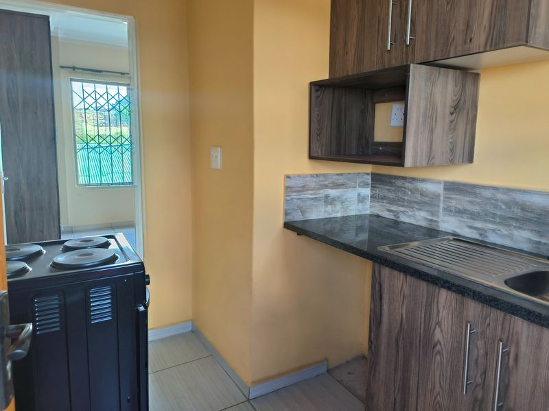 A compact kitchen featuring wood-look cabinetry, dark countertops, a stainless steel sink, and a freestanding stove, with tiled flooring throughout.