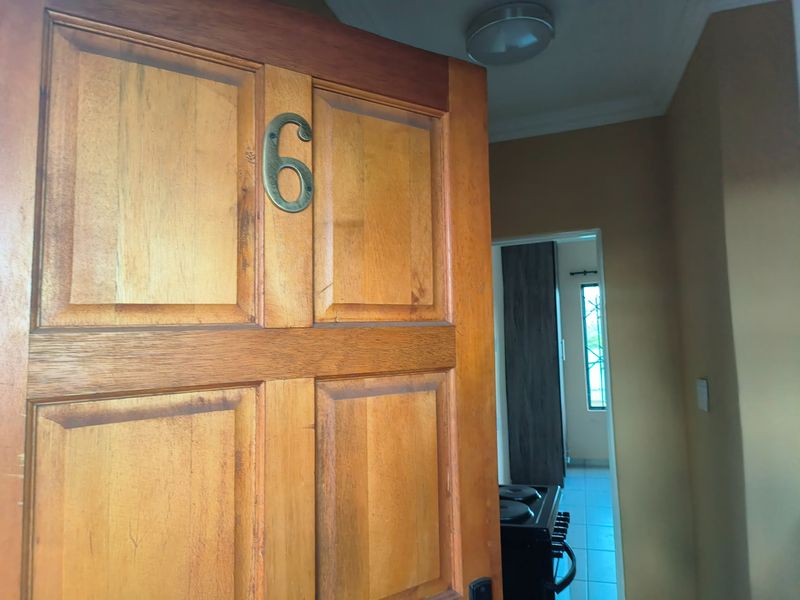 An interior view of a wooden entrance door with a number plate, leading into a hallway with a glimpse of an adjacent room.