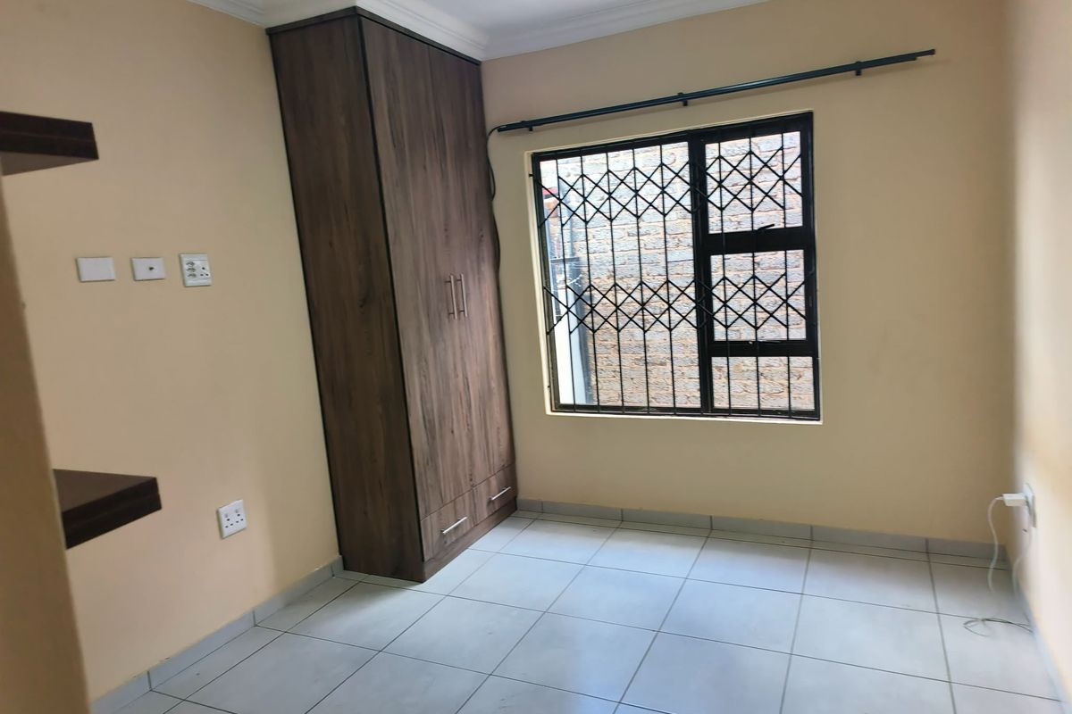 A clean, empty bedroom featuring tiled flooring, a built-in wooden wardrobe, and a window fitted with a security gate.