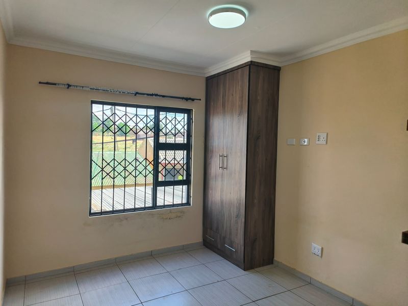 A clean, empty bedroom featuring tiled flooring, a built-in wooden wardrobe, and a window fitted with a security gate.