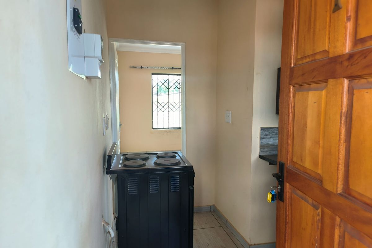 A compact kitchen area featuring tiled flooring, an electric stove, and a wooden entrance door leading to an adjacent room with security bars.