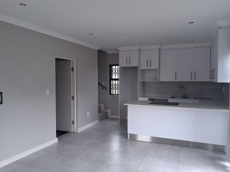 Modern open-plan kitchen featuring white cabinetry, a central island with a hob, and neutral tiled flooring, leading to a staircase and adjacent room.