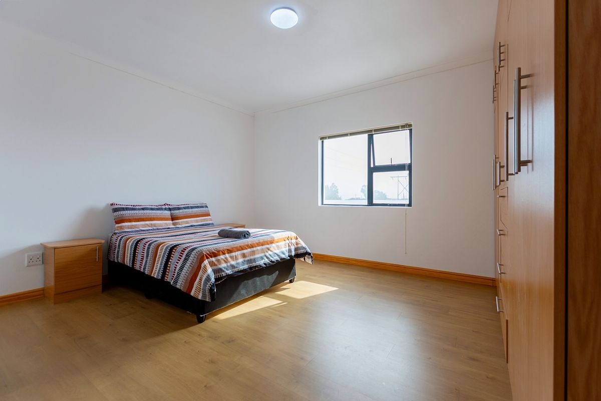 A bright bedroom featuring light wood-look flooring, built-in wooden cupboards, and a large window providing natural light.