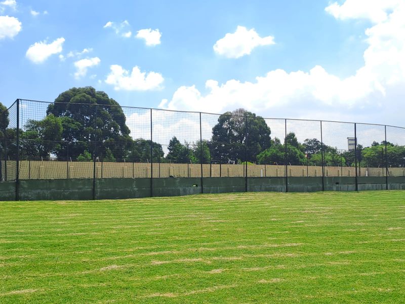 A well-maintained open grass sports field enclosed by high perimeter fencing and a concrete retaining wall.
