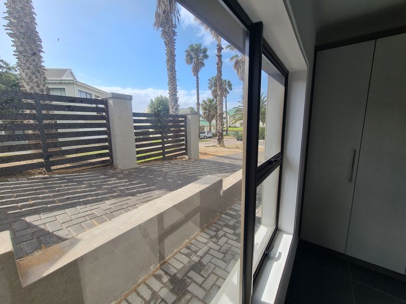 A bright bedroom featuring built-in wardrobes, tiled flooring, and a large window offering a view of the paved outdoor area and garden.
