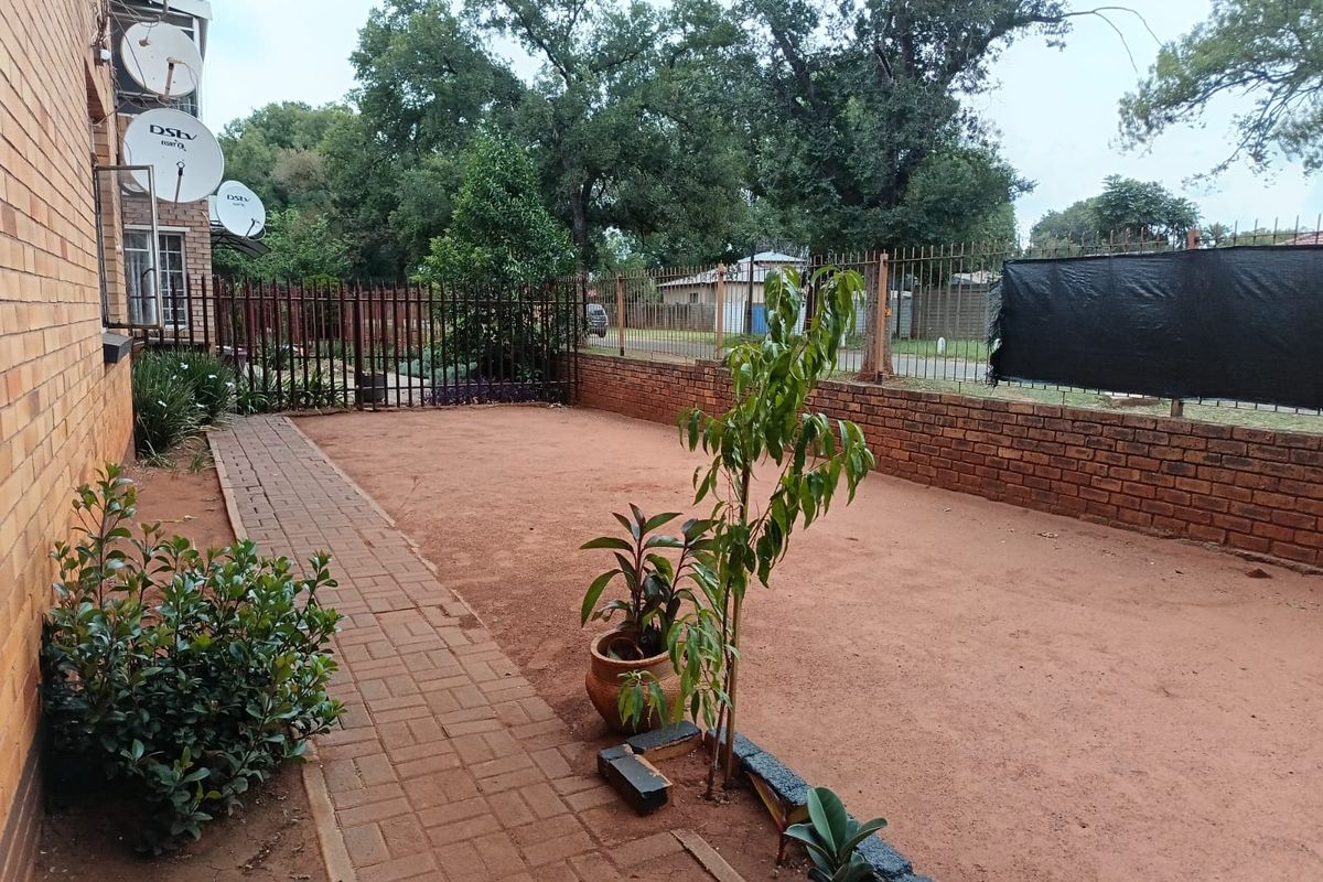A rear exterior area featuring a paved walkway, brick boundary walls, and metal security fencing, currently with an unlandscaped bare earth surface.