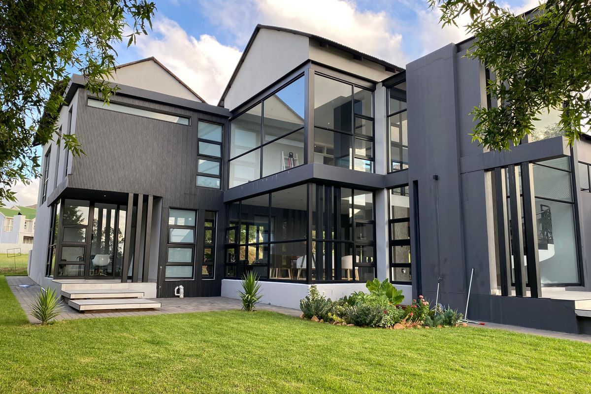 A modern, architecturally designed rear facade featuring extensive glass walls, dark grey cladding, and a well-maintained garden with a paved patio area.