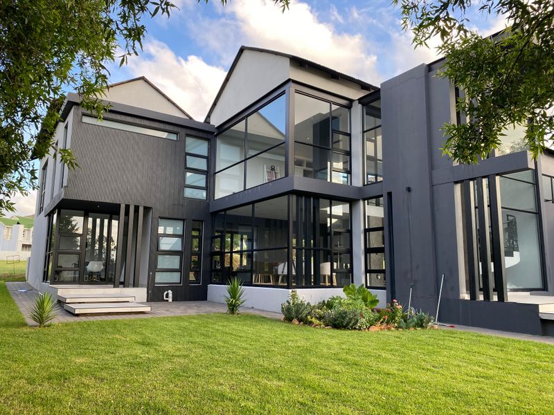 A modern, architecturally designed rear facade featuring extensive glass walls, dark grey cladding, and a well-maintained garden with a paved patio area.