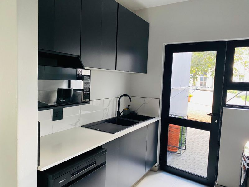 Modern scullery featuring sleek dark cabinetry, stone countertops, marble-look splashback, and direct access to the exterior via a glass-panelled door.