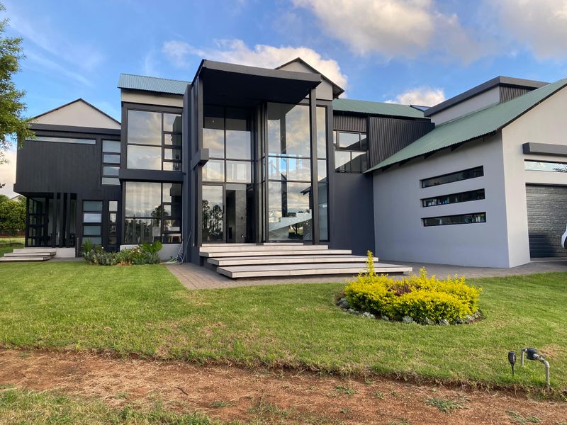Modern architectural home featuring a striking glass facade, dark metal cladding, and a well-maintained front garden with paved entrance steps.