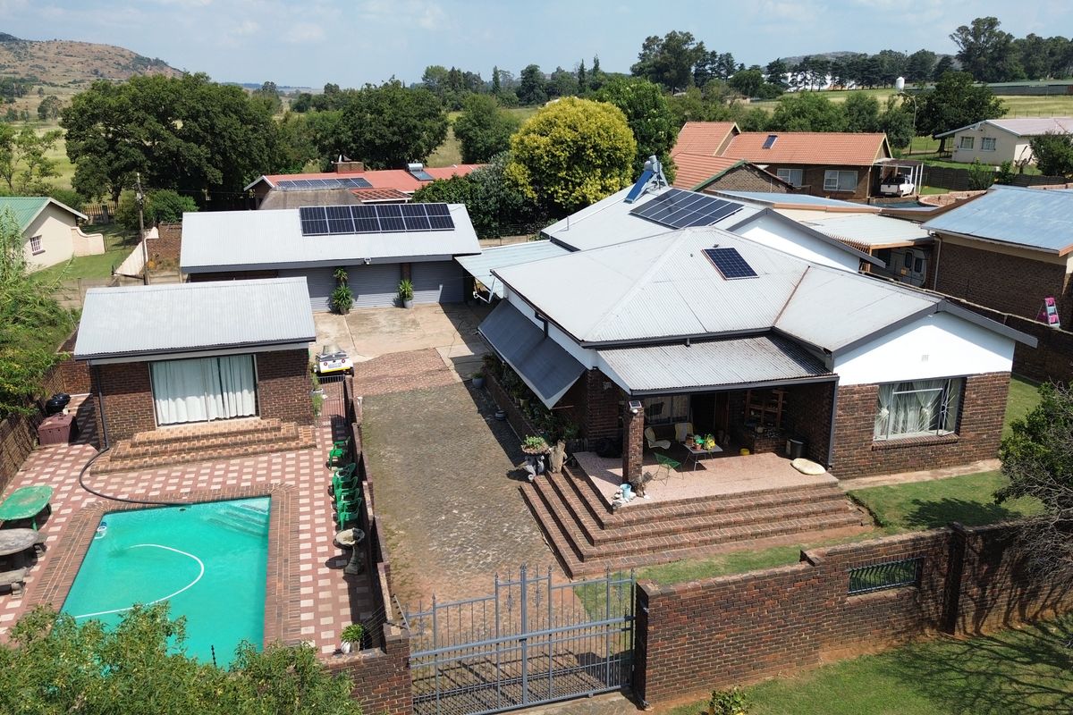 Aerial view of a residential property featuring a swimming pool, multiple outbuildings, solar panel installations, and a paved driveway within a fenced perimeter.