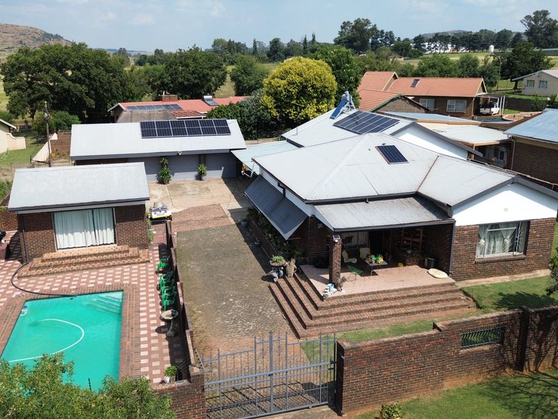 Aerial view of a residential property featuring a swimming pool, multiple outbuildings, solar panel installations, and a paved driveway within a fenced perimeter.