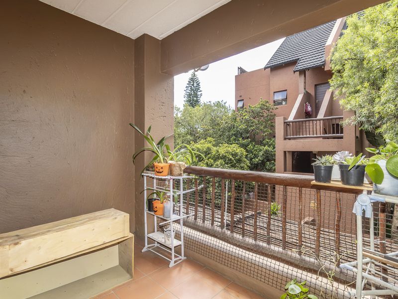 A covered balcony featuring tiled flooring and a timber railing with safety mesh, overlooking the surrounding apartment complex and mature trees.