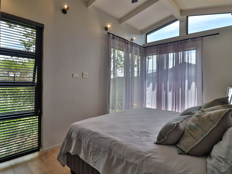 A bright, modern bedroom featuring exposed rafters, clerestory windows, and large glass panels that provide ample natural light and garden views.