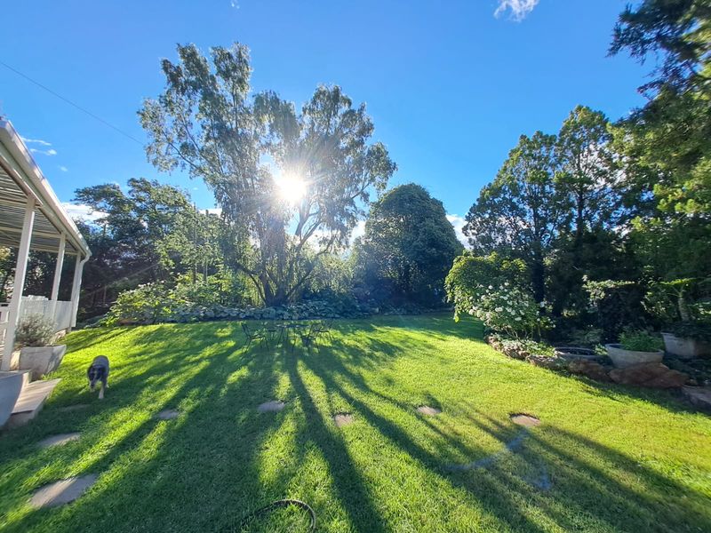 A spacious, well-maintained garden featuring a lush green lawn, mature trees, and stone stepping stones, adjacent to a covered patio area.