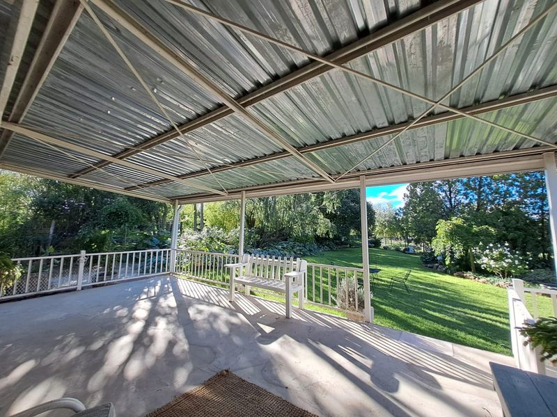 A covered outdoor patio featuring a corrugated metal roof, metal support posts, and a railing, overlooking a spacious green garden.