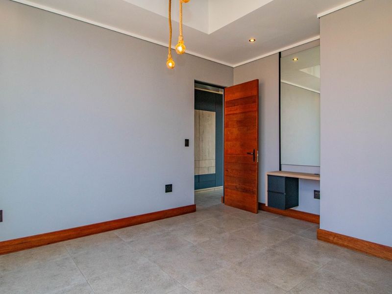 A clean, modern bedroom featuring tiled floors, a built-in vanity with mirror, and a warm wooden door, offering a neutral canvas for personalization.