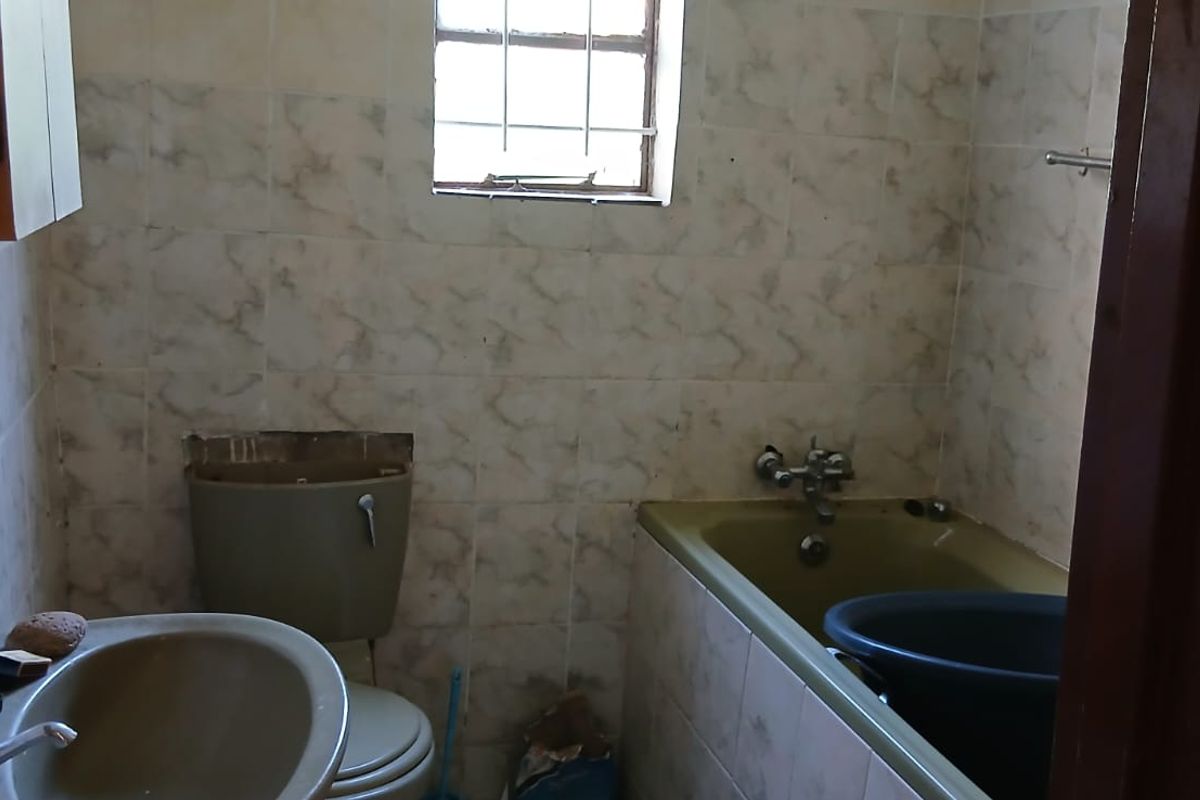 A dated bathroom featuring original fixtures, including a bathtub, toilet, and washbasin, with tiled walls and a window with security bars.