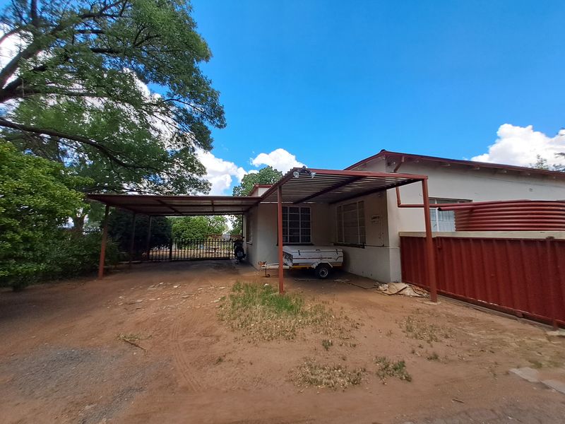 Front exterior view of the property featuring a large metal carport, unpaved driveway area, and perimeter fencing.