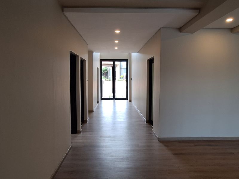 A spacious, well-lit hallway featuring modern laminate flooring, recessed ceiling lights, and a staircase, leading towards a double glass door entrance.