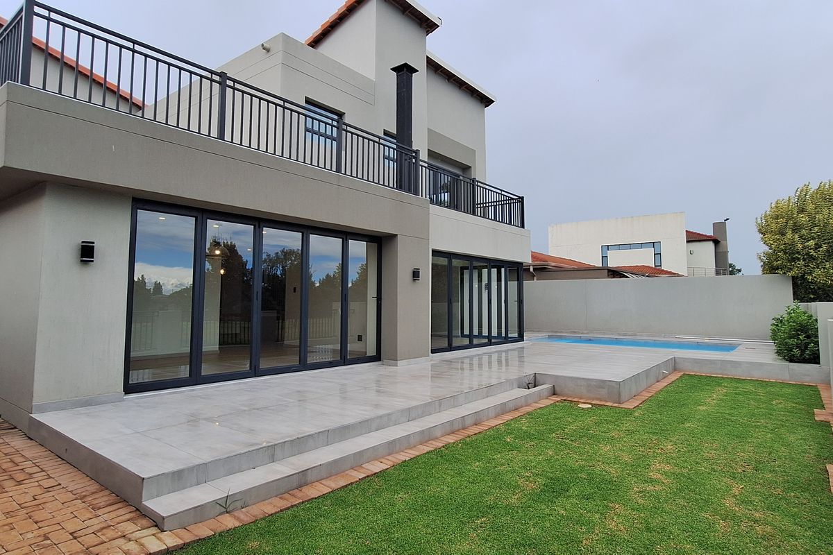 Rear view of a modern double-story house featuring a tiled patio, swimming pool, manicured lawn, and a spacious upper-level balcony with metal railings.