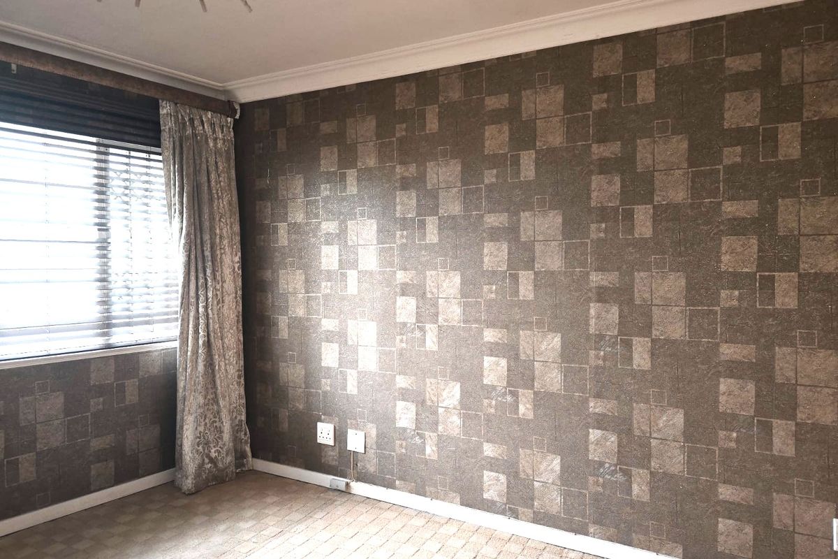 A bedroom featuring patterned wallpaper, carpeted flooring, and a window with fitted blinds and curtains.