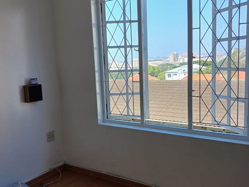 A bright, empty bedroom featuring laminate flooring, security bars on the windows, and a pleasant elevated view of the surrounding neighborhood.