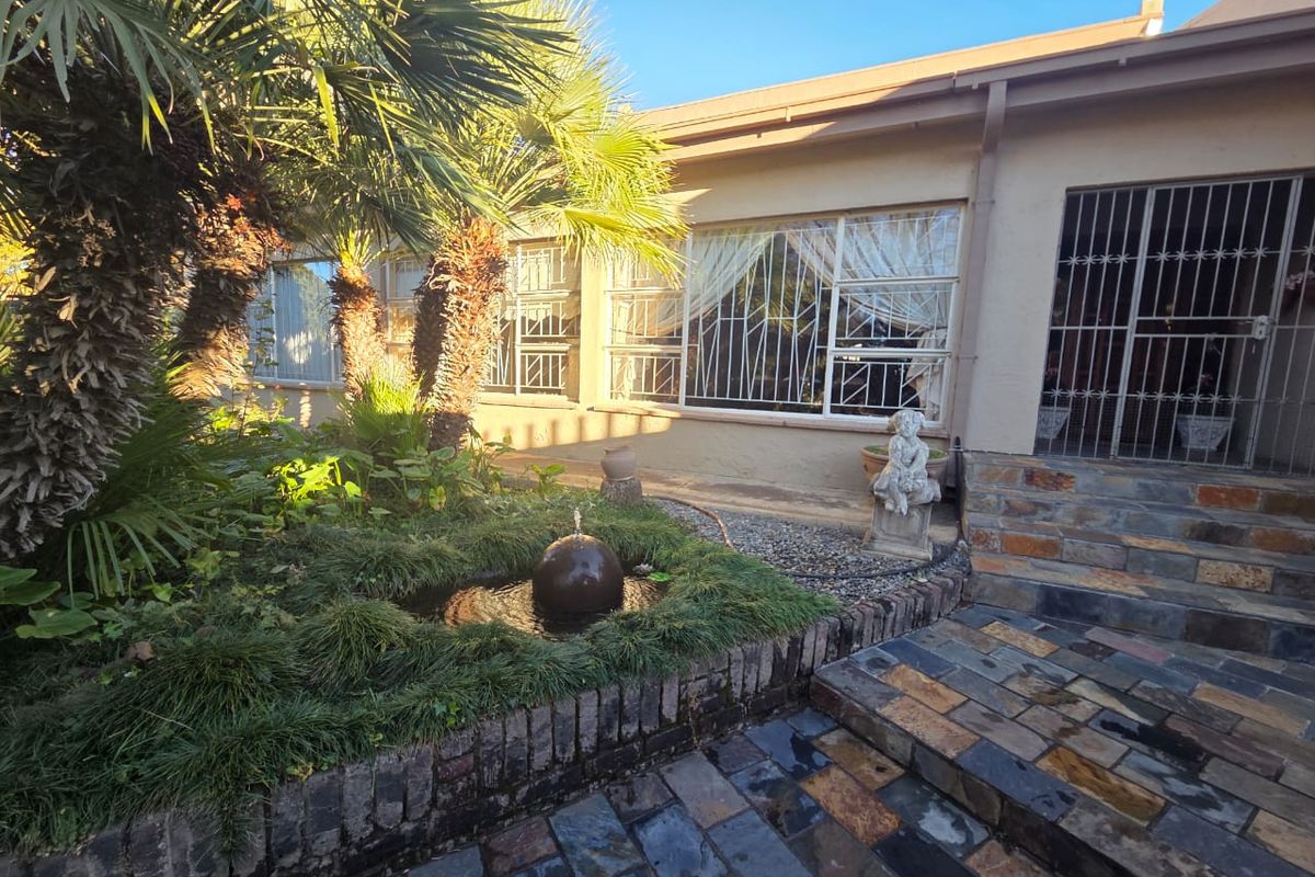 A rear garden area featuring a stone-paved patio, a small water feature, mature palm trees, and security bars on the windows and doors.