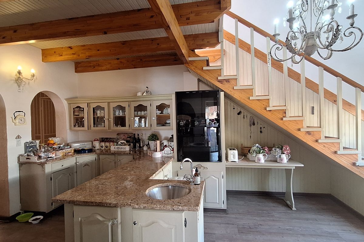 A spacious kitchen featuring a central island with a prep sink, built-in cabinetry, and exposed timber ceiling beams, situated beneath an open staircase.