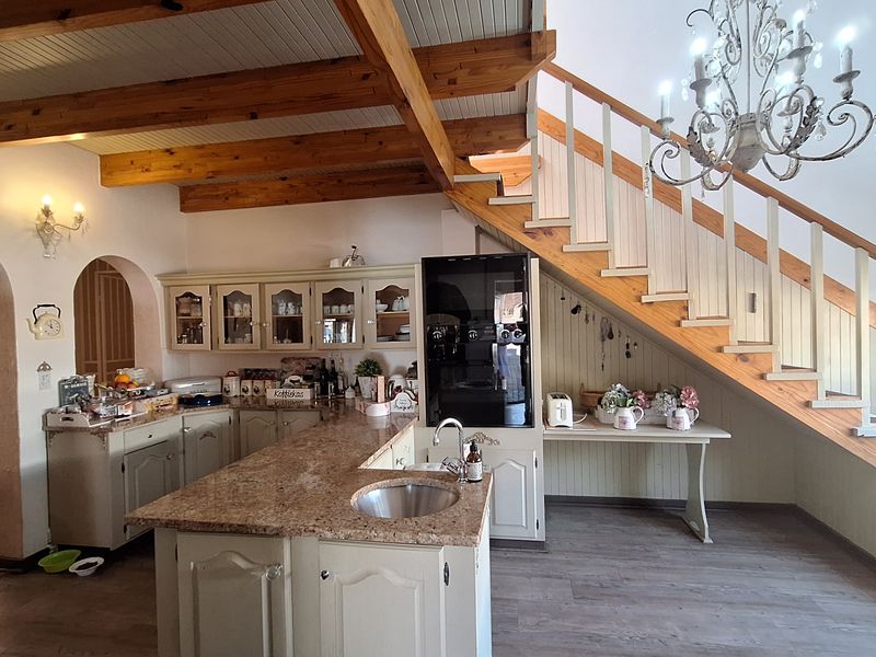 A spacious kitchen featuring a central island with a prep sink, built-in cabinetry, and exposed timber ceiling beams, situated beneath an open staircase.