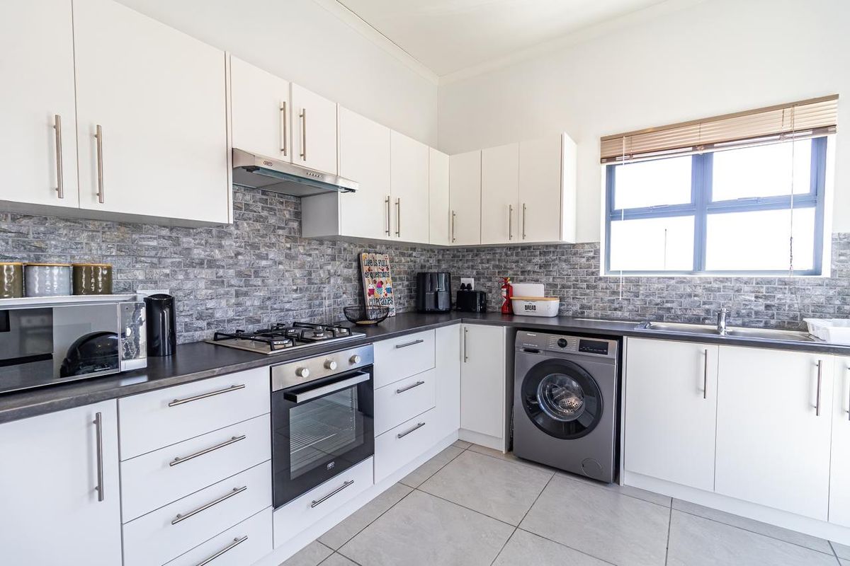 Modern kitchen featuring white cabinetry, dark countertops, a stone-look tiled backsplash, built-in oven, gas hob, extractor fan, and integrated laundry appliance space.