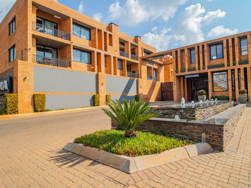 Modern brick apartment complex entrance featuring a paved driveway, water feature, and secure access gates for residents and visitors.