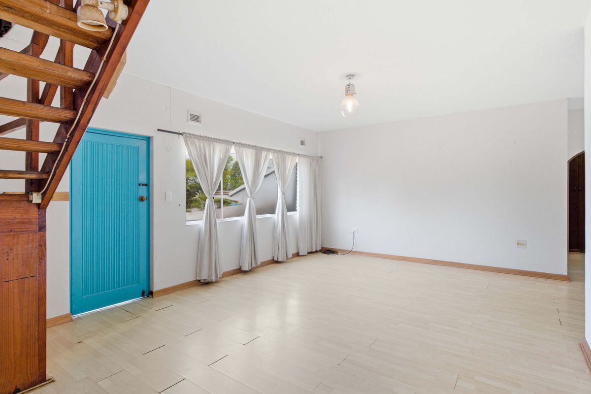 An empty, bright living room featuring light laminate flooring, a wooden staircase, and a vibrant blue entrance door.