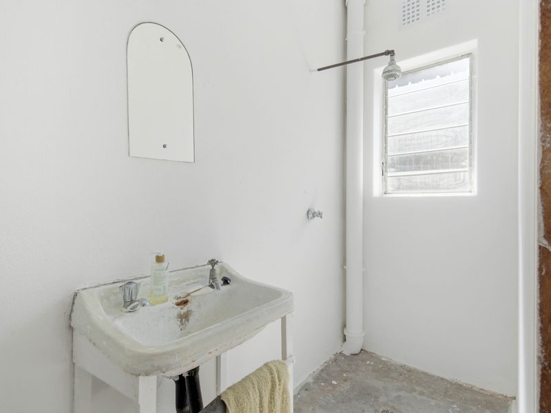 A basic, dated bathroom featuring a wall-mounted basin, exposed shower plumbing, and a louvre window, requiring significant renovation and modernization.