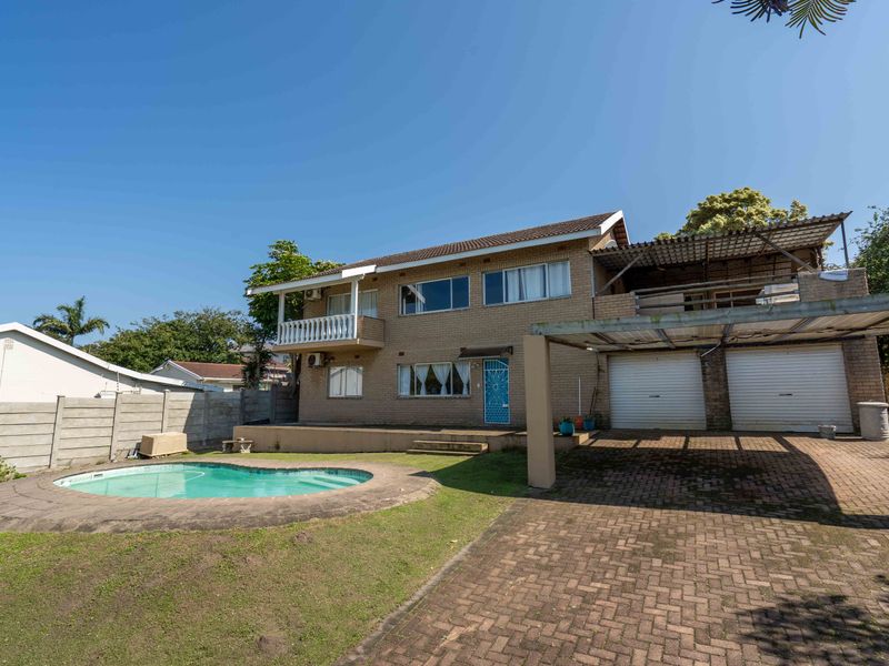 Rear view of a double-story brick house featuring a swimming pool, paved driveway, double garage, and an upper-level balcony.