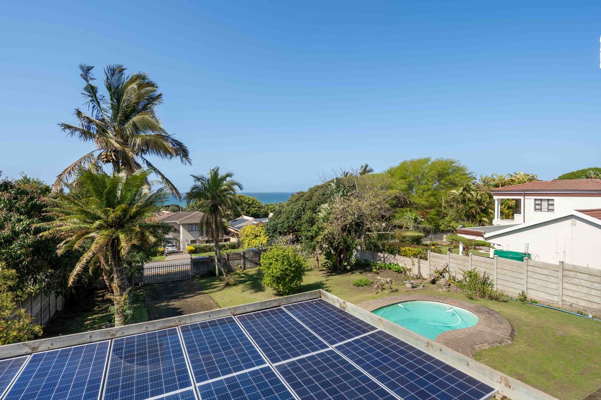 Aerial view of the property grounds featuring a swimming pool, established garden, boundary wall, and roof-mounted solar panel array with distant ocean views.