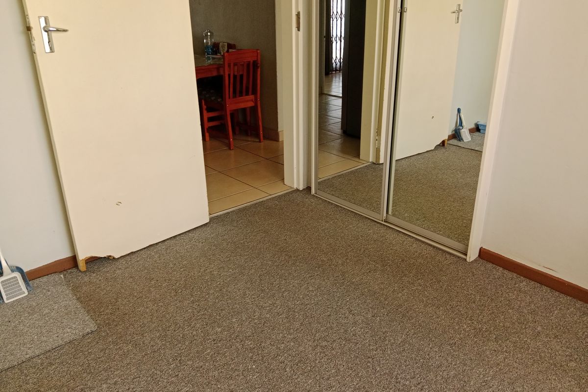 A bedroom interior featuring built-in mirrored cupboards and carpeted flooring, with an adjacent view into a tiled living area.