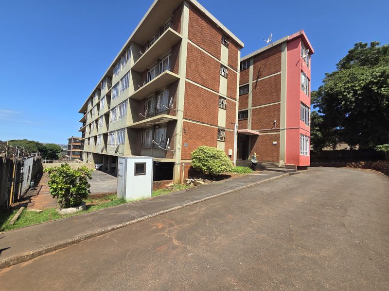 Exterior view of a multi-story brick apartment building featuring concrete balconies, an asphalt driveway, and secure access points.