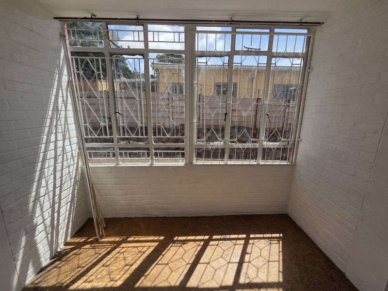 A sunlit, enclosed patio area featuring painted brick walls, large windows with security bars, and worn flooring, offering a blank canvas for renovation.