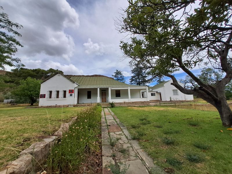 A charming single-story farmhouse featuring a covered veranda, pitched roof, and stone foundation, set within a spacious garden with a stone pathway.