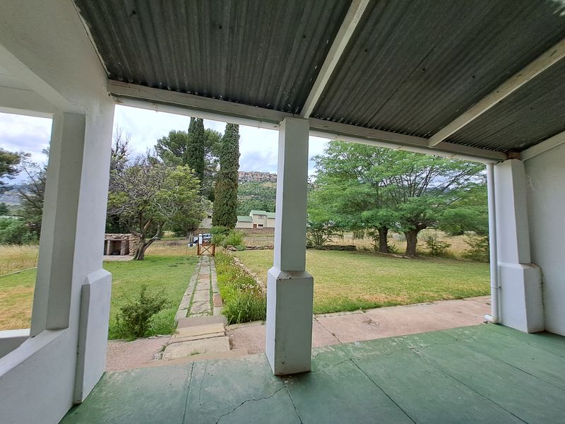 A covered patio area overlooking a spacious garden with a stone pathway, mature trees, and scenic mountain views in the distance.