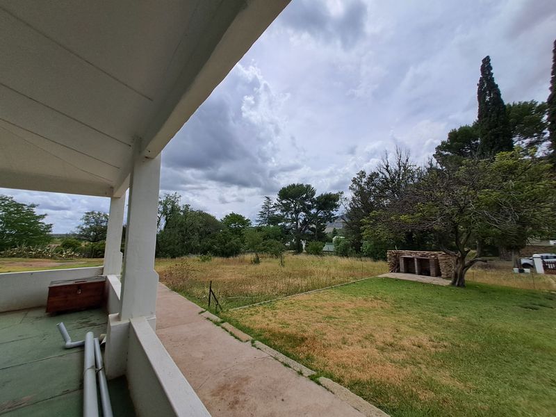 Covered patio overlooking a spacious rear garden featuring a built-in stone braai area and mature trees.