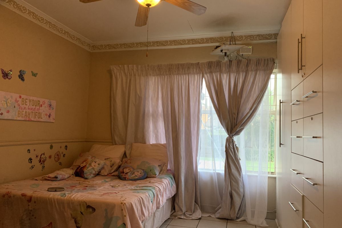 A bedroom featuring built-in cupboards, tiled flooring, and a ceiling fan, with security bars on the window.