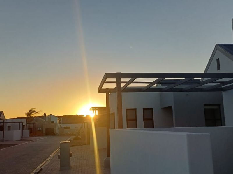 A street-level view of a modern residential development at sunset, featuring clean architectural lines, paved driveways, and contemporary metal pergola structures.