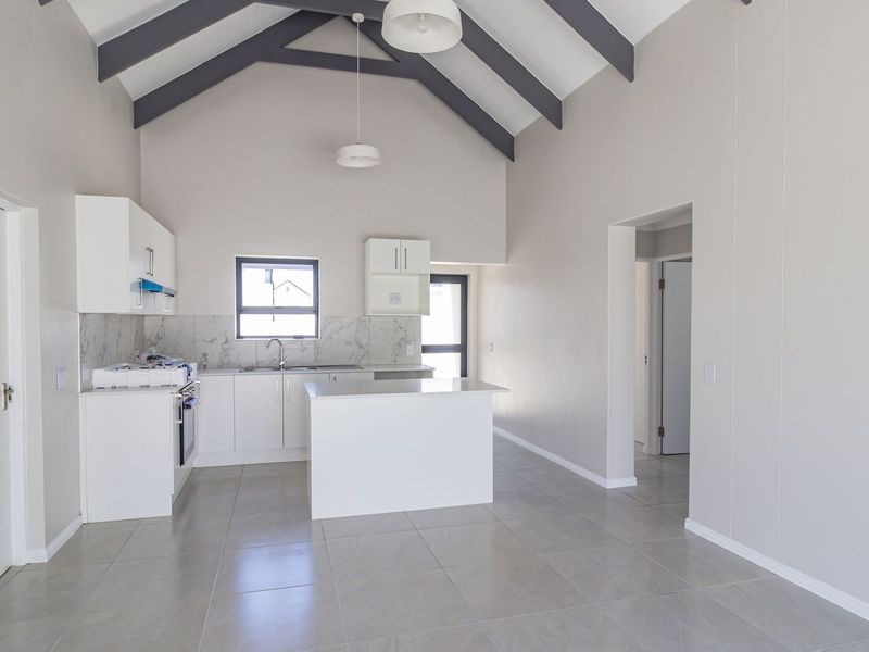 Bright open-plan kitchen and living area featuring a high vaulted ceiling with exposed rafters, modern white cabinetry, stone backsplash, and neutral tile flooring.