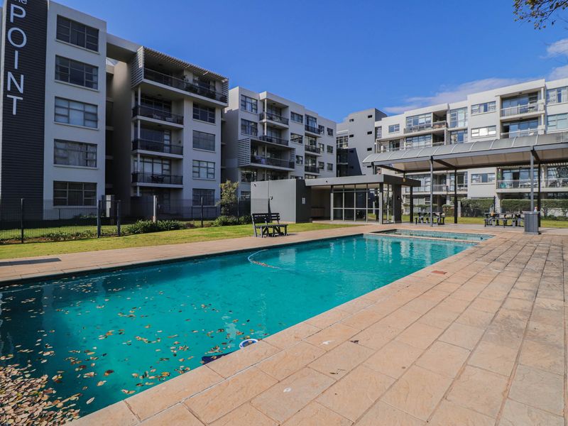 Communal swimming pool area featuring a paved deck, covered patio structure, and landscaped garden within a modern apartment complex.