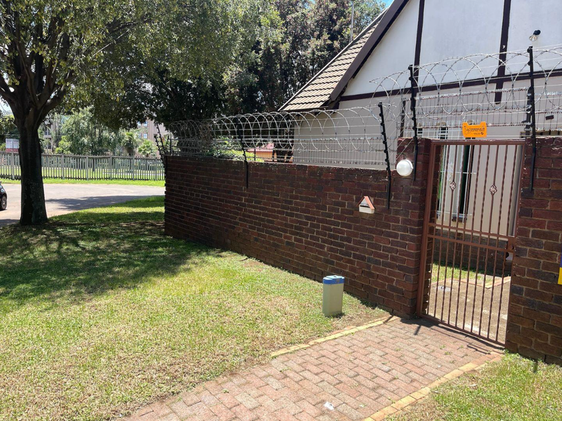Front exterior view featuring a brick boundary wall with integrated electric fencing and razor wire, a metal pedestrian gate, and a paved walkway.