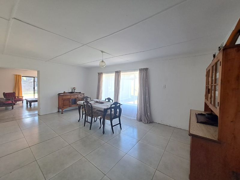 A spacious dining area featuring large format floor tiles, neutral walls, and a ceiling with visible panel joints, leading into an adjacent living space.